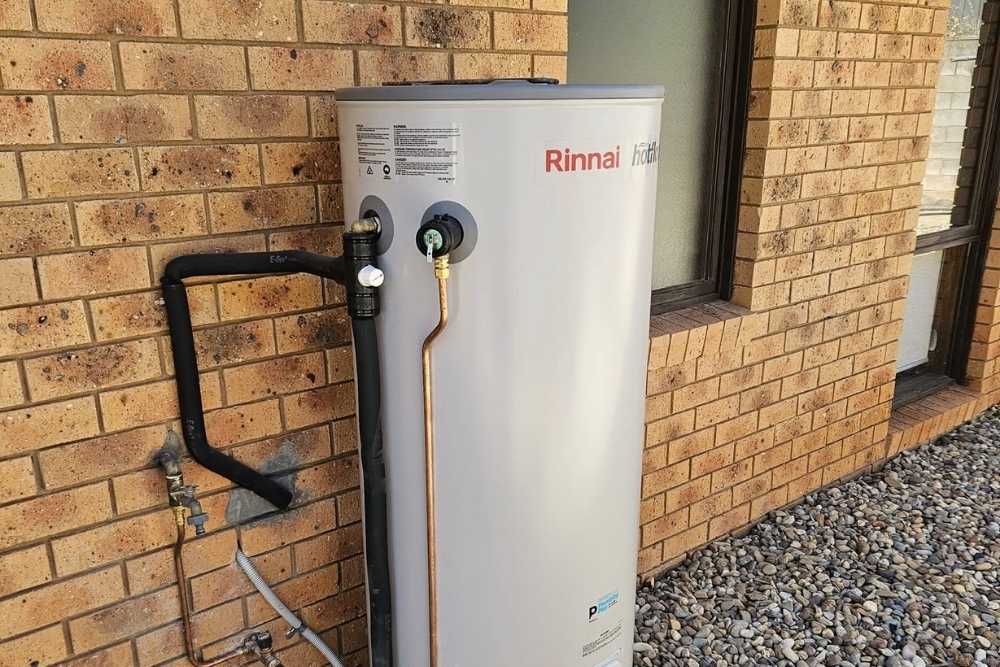 A grey hot water system tank installed outside against a brick wall with neat copper plumbing pipes-